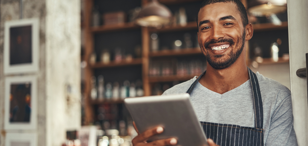 Smiling male business owner holding a digital tablet while standing in the doorway of his cafe